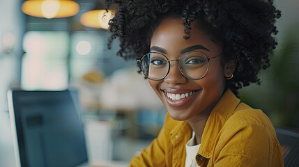 Obraz premium A woman with curly hair and glasses is smiling at the camera. She is wearing a yellow shirt and sitting in front of a computer