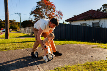 Little boy riding trike on driveway of home on sunny day dad helping