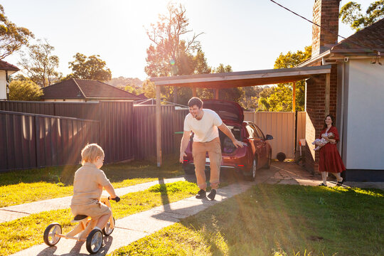 Family of four packing car for holidays with toddler riding on trike in golden sunlight