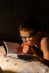 Close-up of a child reading in the dark by lamp light. A child with poor eyesight wearing glasses...