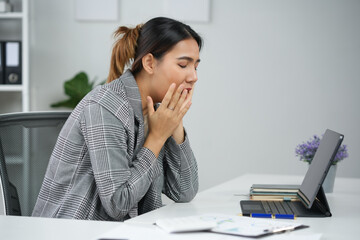 Tired businesswoman yawning at her desk while working on a tablet. Work fatigue and workplace burnout concept.