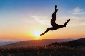 Fototapeta premium Silhouette of male ballet dancer jumping and doing a majestic dance move in nature, Behind him are heavenly clouds and the sun is setting.