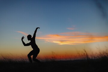 Silhouette of male ballet dancer doing a majestic dance move in nature, Behind him are heavenly clouds and the sun is setting.