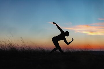 Silhouette of male ballet dancer doing a majestic dance move in nature, Behind him are heavenly clouds and the sun is setting. © Zenstratus