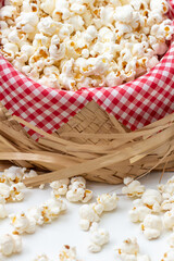 Centered close-up of the front view of a tilted straw hat with red and white checkered fabric and salted popcorn inside. With loose popcorn spread across the front.