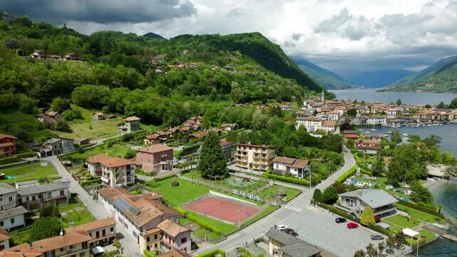 An aerial shot captures a picturesque town nestled by a lake with lush greenery and clouds overhead. Pella, Piedmont. Orbit Motion Shot