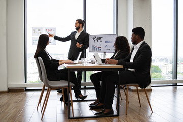Confident business man giving presentation to multiracial colleagues standing near graphs and charts on window glass. Professional discussion about work strategies and statistics.