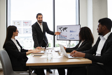 Businessman presenting to colleagues in modern meeting room. Group discussing data and statistics displayed on screen and charts. Professional team engaged in business discussion around table.