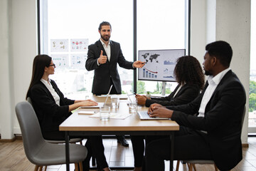 Businessman reports to colleagues standing near graphs and charts on monitor, screen, display. Team engaged in corporate meeting discussing sales, performance, and strategies.