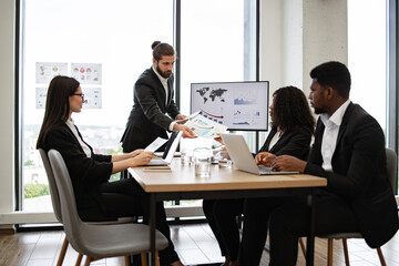 Colleagues working on laptops, analyzing charts and graphs in modern office setting Businessman hands out paper tasks or results of company's activities to colleagues during meeting.