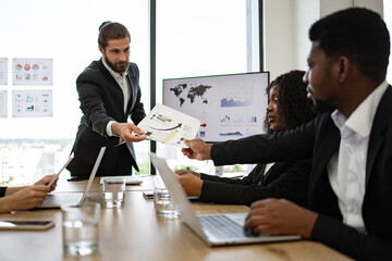 Businessman handing out paper tasks or results of company activities to colleagues during a meeting. Teamwork and collaboration in modern office setting with digital devices and documents.