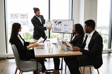 Business team in meeting room discussing financial presentation with charts. Colleagues in professional attire collaborating on project. Office setting with large windows and natural light.