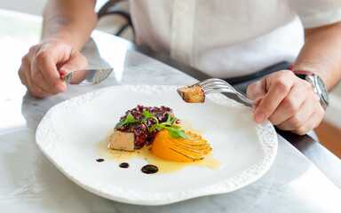 Hands of young man with knife and fork cutting piece of Foie Gras on white plate while having lunch  by served table