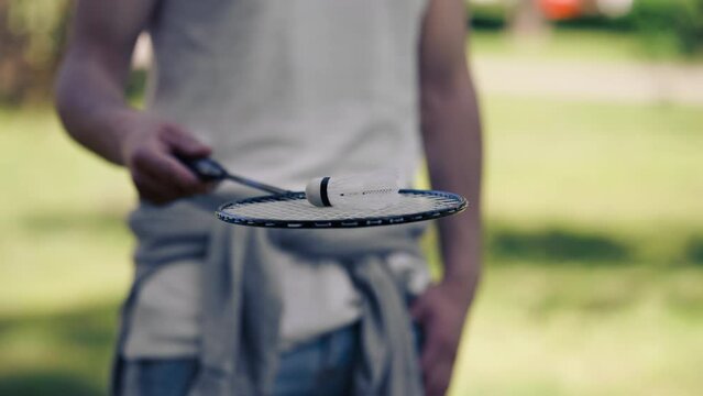 outdoors in a park with trees young handsome brunette guy in jeans and a white T-shirt, a brunette stuffing a shuttlecock warming up before playing bambinton