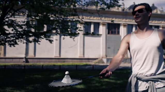 outdoors in a park with trees young handsome brunette guy in jeans and a white T-shirt, a brunette stuffing a shuttlecock warming up before playing bambinton