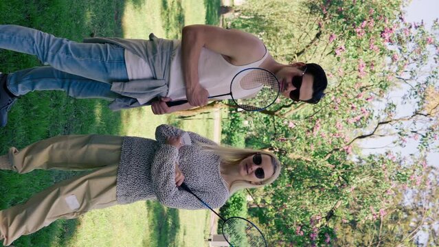 vertical video outdoors in a park with trees young brunette guy stands next to a blonde girl bambinton rackets best friends relaxing together