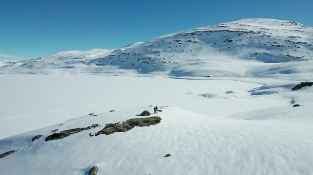 Two hikers standing next to a tent on the snowy landscape of the frozen Tyin Lake in Norway, with mountains in the background.