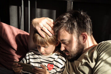 Obraz premium Son and dad are having a great time together. They are looking at the smartphone together and hugging. Concept of a happy childhood and parenthood.