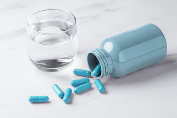 Blue pill capsules spilling out of bottle and glass of water on white table background