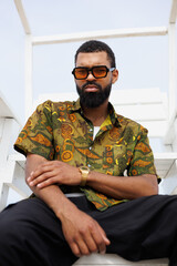 Young african american man in streetwear and sunglasses sitting on lifeguard tower on beach