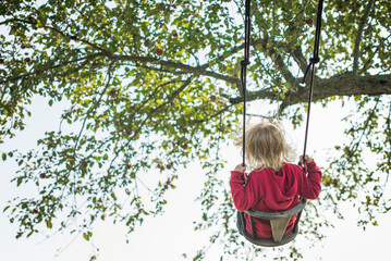 Child in a red hoodie swinging under a tree with green leaves