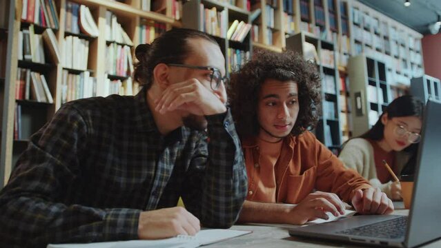 Young middle-eastern college student discussing homework on laptop with group mate at desk in public library