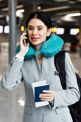 Business woman with tickets, passport , backpack and neck pillow talking on phone at airport lobby....