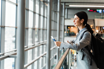 Tourist in airport is standing near panoramic window waiting for security check. Woman holding passport, boarding pass and smartphone standing near window at airport lobby and waiting for departure.