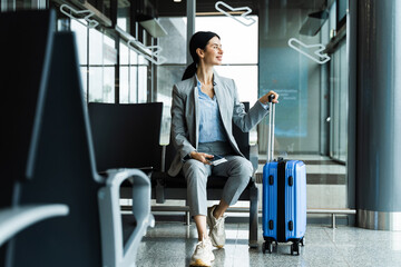 Woman is holding handle of blue suitcase and looking around airport terminal. Business woman sitting in airport and waiting for starting her travel voyage.