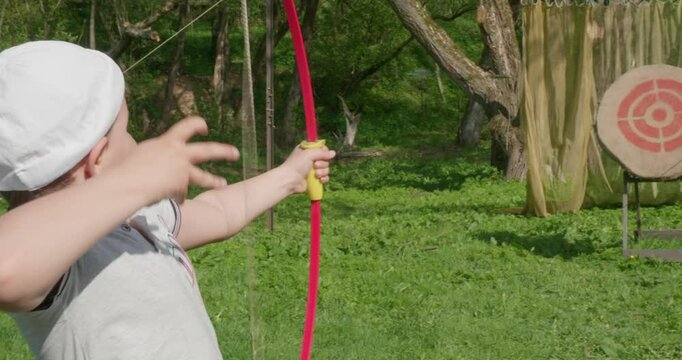Having taken aim, the child pulls the bowstring and shoots the arrow right at the target. A child shoots a bow at a shooting range in a park while walking. The child is interested in archery.