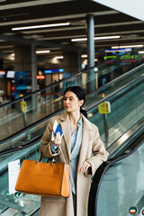 Traveler holding boarding pass, passport, smartphone and handbag while standing near escalator in airport terminal. A woman in a coat walks to the boarding gate