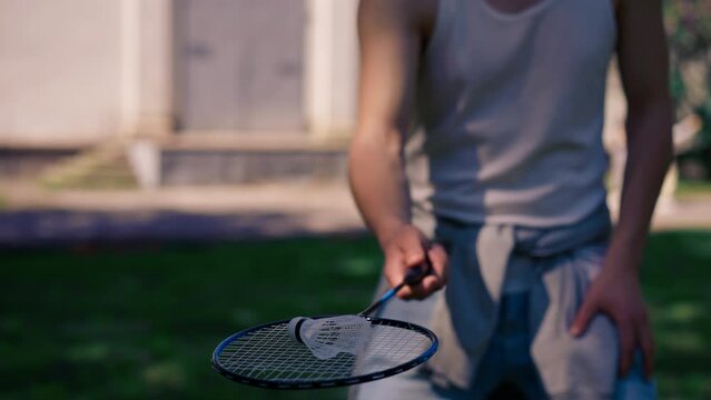 outdoors in a park with trees young handsome brunette guy in jeans and a white T-shirt, a brunette stuffing a shuttlecock warming up before playing bambinton
