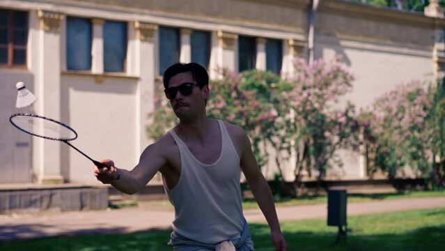 outdoors in a park with trees young handsome brunette guy in jeans and a white T-shirt, a brunette stuffing a shuttlecock warming up before playing bambinton