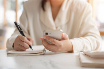 Fototapeta premium A cropped image of a woman writing something in her notebook and using her smartphone.