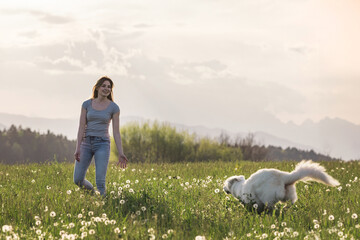 Young woman playing with their pet dog on a spring meadow at sunset with mountains in the background.