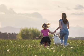 Idyllic scene of mother, daughter and pet dog happily running in the grass field full of...