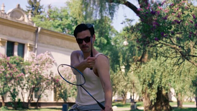 on the street in the park among the trees young brunette guy in a white T-shirt is fooling around with two rackets for playing bambinton