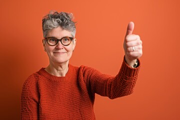 Portrait of a tender woman in her 50s showing a thumb up while standing against solid color backdrop