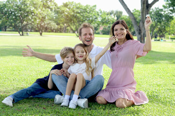 Fototapeta premium Happy family sitting on green grass and waving hands at park, smiling father, mother, son and daughter spending time together outdoors in beautiful garden on holiday. Parents and kids have fun outside