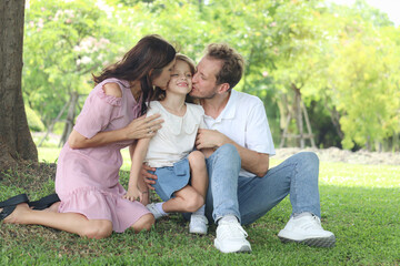 Fototapeta premium Happy beautiful lovely family in outside garden. Joyful parents kissing their daughter girl while sitting under tree at summer green park. Father, mother, and kid child spending time together outdoor.