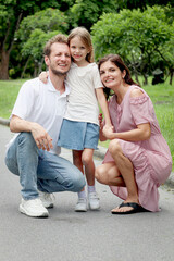 Fototapeta premium Portrait of happy beautiful family in outside garden. Smiling daughter girl hugging parents on park walkway at summer green park. Joyful father, mother, and kid child spending time together outdoor.