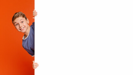 Cute smiling boy looking out of white blank placard ready for your text or product, orange background