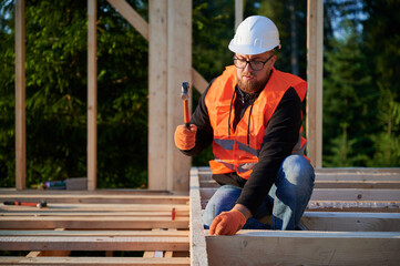 Wooden frame, two-story abode is being built by carpenter near forest. Bearded man wearing glasses is driving nails with a hammer, attired in a safety helmet and a construction vest.