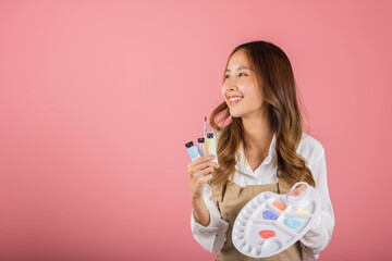 Asian beautiful young woman artist holding brush and paint palette, Happy female painting using paintbrush and palette with colors, studio shot isolated on pink background, Paintings and art equipment