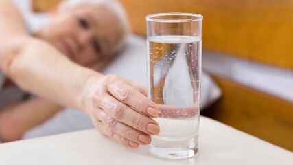 Thirsty senior woman taking glass of water lying in bed, start new day with fresh aqua, panorama