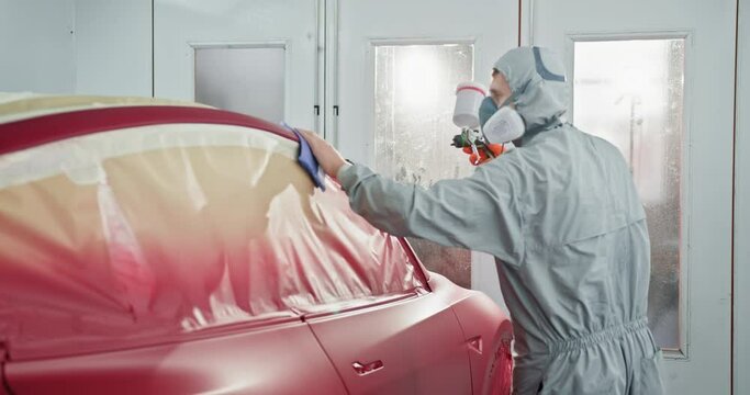 Technician in protective gear spray painting a red car part in a specialized chamber at a car service station. Car Painting Technician Applying Red Paint in Auto Body Shop. Car manufacturing factory.