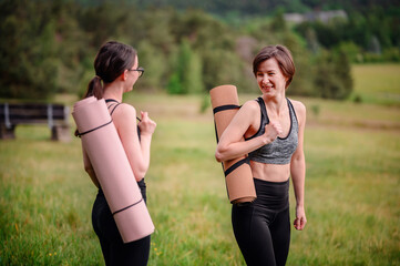 Two women in athletic wear walk through a grassy field, smiling and carrying rolled yoga mats on their shoulders, ready for a yoga session in nature.