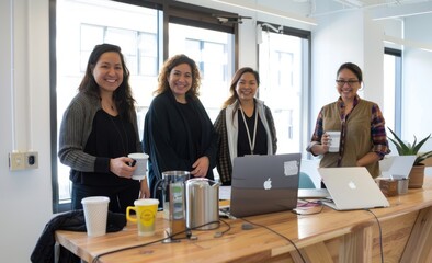 Obraz premium Four diverse women in their thirties standing behind an office desk, smiling at the camera while holding coffee mugs and laptops