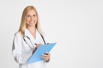 A smiling lady doctor in a white coat is writing on a blue clipboard while holding a stethoscope. The doctor is standing in front of a plain white background, copy space