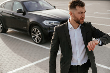 Checks the time. Businessman in suit is near his black car outdoors